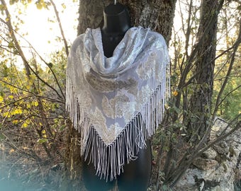 May include: A light gray scarf with a floral pattern and white fringe. The scarf is draped over a black mannequin. The background includes trees and sunlight.