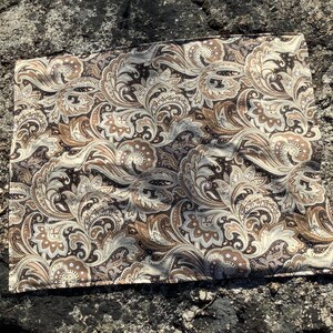 May include: A rectangular fabric piece featuring a dense, intricate paisley pattern in shades of dark brown, tan, and cream. The design consists of swirling, teardrop-shaped motifs and floral elements that repeat across the entire surface of the textile.
