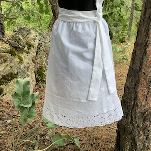 May include: A white apron featuring a wrap-around waist tie and a gathered skirt. The fabric is adorned with small embroidered floral motifs throughout, while the bottom hem displays a wide, intricate eyelet lace border with circular patterns.