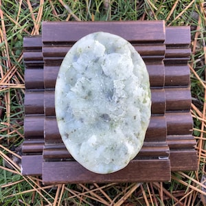 May include: A light green, oval-shaped stone with a textured surface, resting on a dark brown wooden soap dish. The stone has a mix of light and dark green hues with some darker spots. The soap dish has a grooved design.