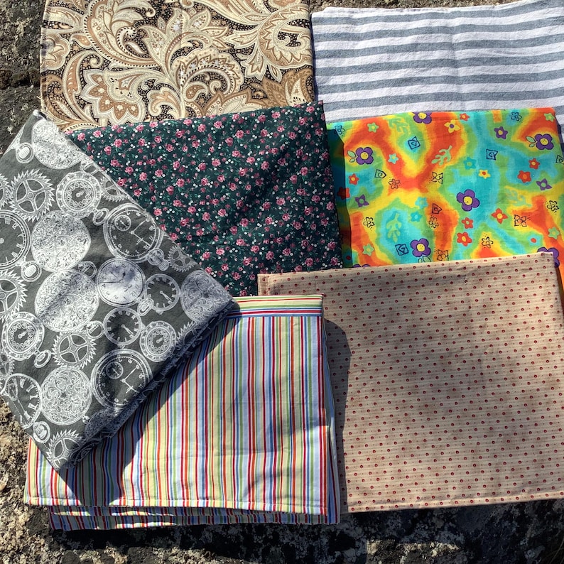 May include: A collection of seven folded fabric squares featuring diverse patterns including a brown paisley print, grey and white stripes, a dark green floral print, a bright multi-coloured psychedelic floral design, a clock gear print, vertical stripes, and tan polka dots.