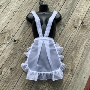 May include: A white, sheer apron with ruffled edges and suspender straps, displayed on a black mannequin. The apron has a classic design, with a gathered skirt and a bib-like top. The background is a weathered wooden surface.