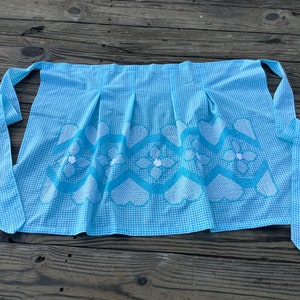May include: A light blue and white gingham apron with a decorative floral and heart pattern. The apron has pleats and long ties for securing around the waist. The apron is made of fabric.
