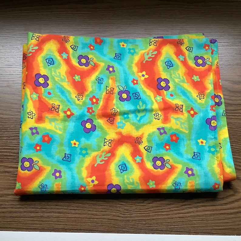 May include: A square fabric piece featuring a vibrant tie-dye background in shades of orange, yellow, and turquoise. The pattern is decorated with small, repeating illustrations of purple, red, and yellow flowers along with green leaf shapes.