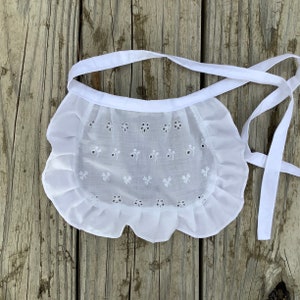 May include: White cotton apron with eyelet lace trim and a ruffled edge. The apron has a simple design with small floral embroidery.
