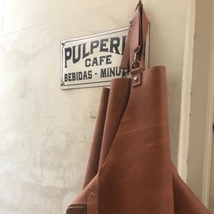 May include: A brown leather apron with a large pocket and adjustable straps. The apron is hanging on a wall with a sign that reads "Pulperia Cafe Bebidas - Minutos".