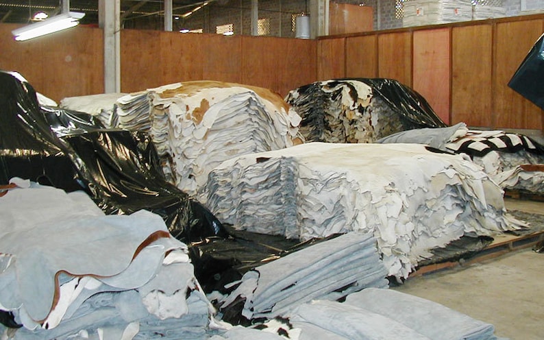 May include: A large warehouse space filled with stacks of tanned animal hides. The hides are stacked in piles and covered with black plastic sheeting. Some of the hides are white, while others are brown and black.
