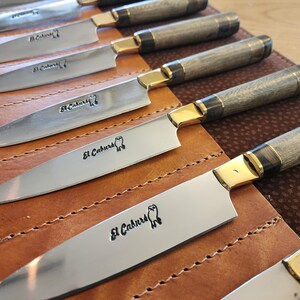 May include: A set of six silver knives with wooden handles and gold accents. The knives are arranged in a leather roll with the brand name "El Caburero" engraved on each blade.