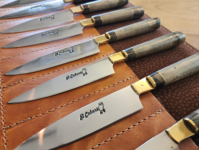 May include: A set of six silver knives with wooden handles and gold accents. The knives are arranged in a leather roll with the brand name "El Caburero" engraved on each blade.