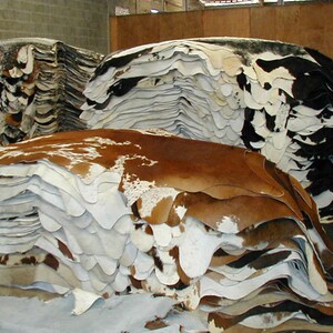 May include: A large stack of cowhide rugs in various shades of brown, white, and black. The hides are stacked in a warehouse setting.