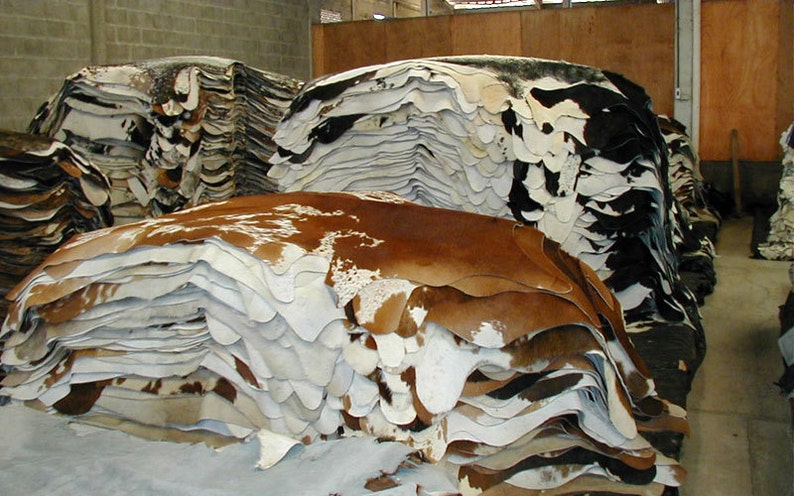 May include: A large stack of cowhide rugs in various shades of brown, white, and black. The hides are stacked in a warehouse setting.