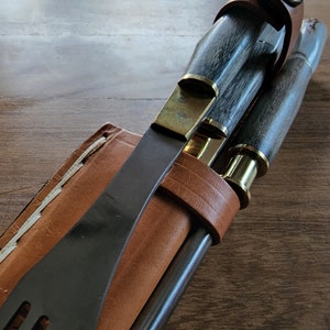 Puede incluir: Una funda de cuero con un cuchillo negro, un cuchillo con mango de madera y lat&oacute;n, y una herramienta con mango de madera y lat&oacute;n. La funda es marr&oacute;n con costuras blancas.