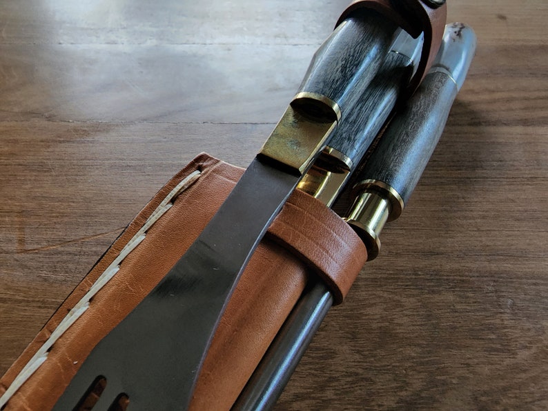 Puede incluir: Una funda de cuero con un cuchillo negro, un cuchillo con mango de madera y lat&oacute;n, y una herramienta con mango de madera y lat&oacute;n. La funda es marr&oacute;n con costuras blancas.