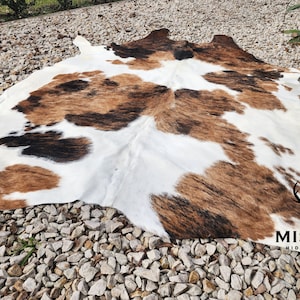 Puede incluir: Una alfombra de piel de vaca marrón, negra y blanca con fondo blanco y manchas marrones y negras. La alfombra está colocada sobre un lecho de pequeñas piedras grises.