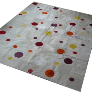Puede incluir: Alfombra de cuero blanco con un diseño de patchwork que presenta círculos coloridos en tonos rojos, naranjas, amarillos y morados.