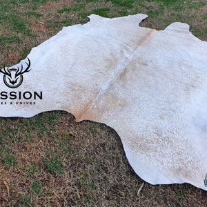 May include: White cowhide rug with brown markings. The rug is laid out on a grassy surface. The text "MISSION HIDES & KNIVES" is visible in the image. The text "EXPORT QUALITY" is visible in a circular stamp.