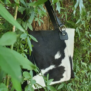 May include: A black and white cowhide tote bag with a black leather strap and tassel. The bag is hanging from a tree branch.