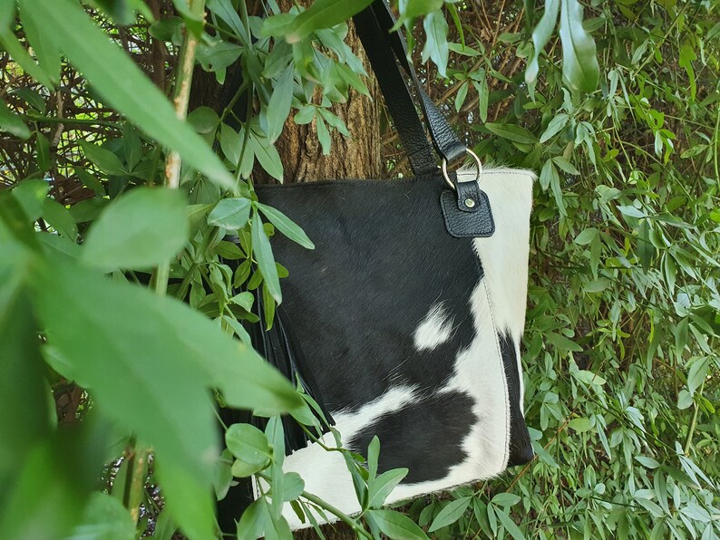 May include: A black and white cowhide tote bag with a black leather strap and tassel. The bag is hanging from a tree branch.