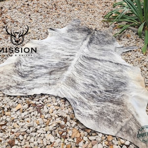 Puede incluir: Una alfombra de piel de vaca gris y blanca con marcas negras. La alfombra está colocada sobre un lecho de pequeñas piedras de grava. El texto "MISSION HIDES & KNIVES" es visible en la esquina superior izquierda de la imagen. El texto "EXPORT QUALITY" es visible en la esquina inferior derecha de la imagen.