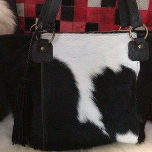 May include: A black and white cowhide tote bag with a fringe tassel. The bag has black leather straps and gold hardware.