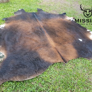 Puede incluir: Una alfombra de piel de vaca marrón y negra con manchas blancas. La alfombra está colocada sobre una superficie de hierba verde. El texto "MISSION HIDES & KNIVES" es visible en la esquina superior derecha de la imagen.