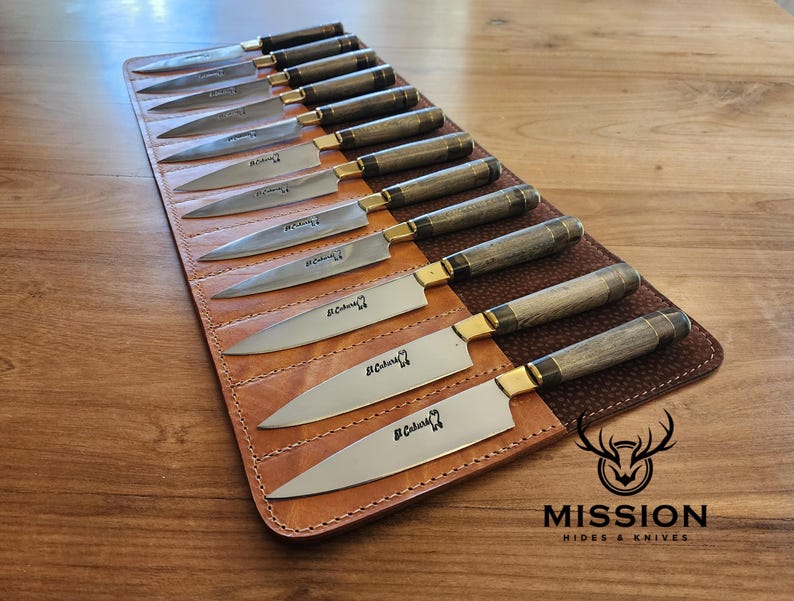 May include: A brown leather knife roll with 12 slots for knives. The roll is open and the knives are visible. The knives have wooden handles and are made by Mission Hides & Knives.