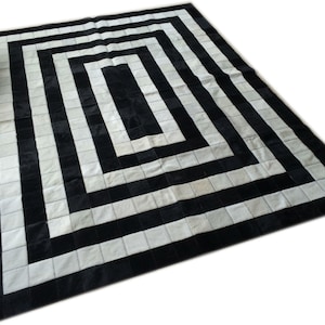 May include: Black and white cowhide rug with a geometric pattern. The rug is made of squares of black and white leather, arranged in a concentric square pattern.