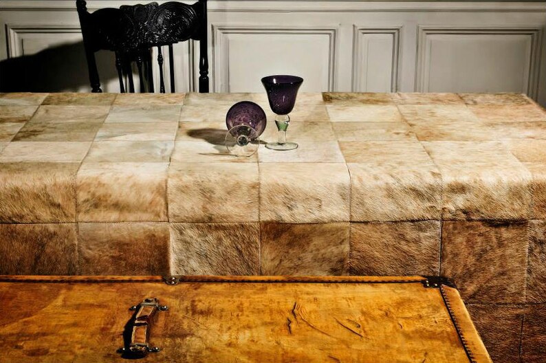 May include: A brown leather table cloth with a patchwork design. Two clear glass wine glasses with purple stems are on the table. A wooden chest with a brass latch is in the foreground.