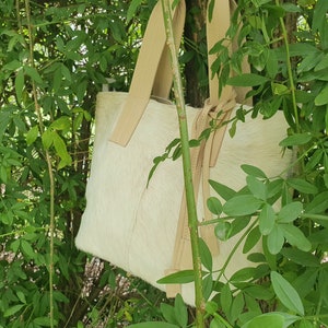 May include: A white leather tote bag with tan leather straps and a knotted detail. The bag is hanging from a branch in a green leafy bush.