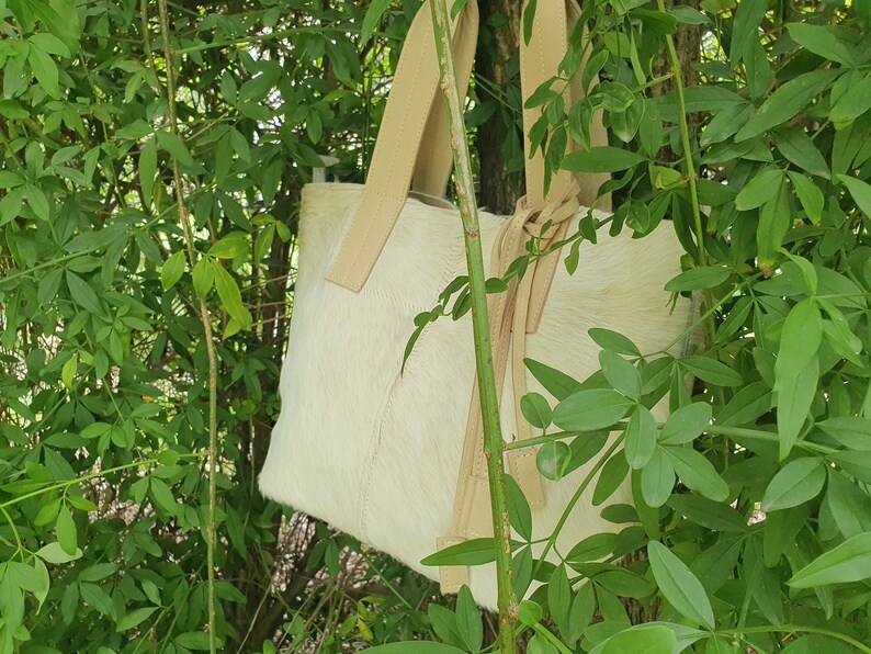 May include: A white leather tote bag with tan leather straps and a knotted detail. The bag is hanging from a branch in a green leafy bush.