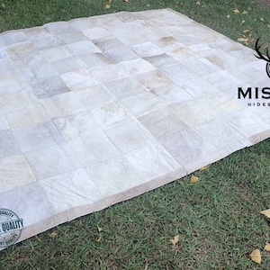 May include: A white cowhide patchwork rug with a tan border. The rug is laid out on a green grassy surface. The text "MISSION HIDES & KNIVES" is visible in the upper right corner of the image. A green and white "EXPORT QUALITY" stamp is visible in the lower left corner of the image.