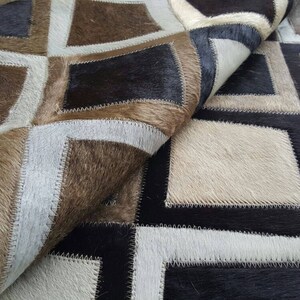 Pode incluir: Um tapete de patchwork feito de couro de vaca em tons de marrom, preto e branco. O tapete é feito de quadrados e retângulos, e as bordas são costuradas juntas.