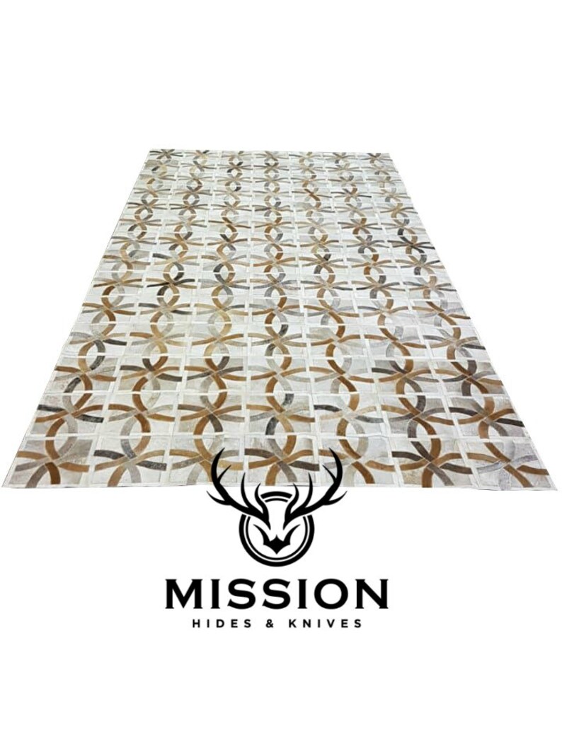 May include: A white leather rug with a geometric pattern of brown and gray circles and ovals. The rug is made of cowhide and has a unique, handcrafted look. Mission Hides & Knives is printed at the bottom of the image.