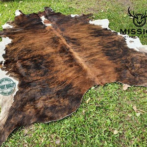 May include: A brown and white cowhide rug with a black and brown pattern. The rug is laid out on a green grassy surface. The text "EXPORT QUALITY" is printed on a white circle with a green border. The text "MISSION HIDES & LEATHERS" is printed in black on a white background.