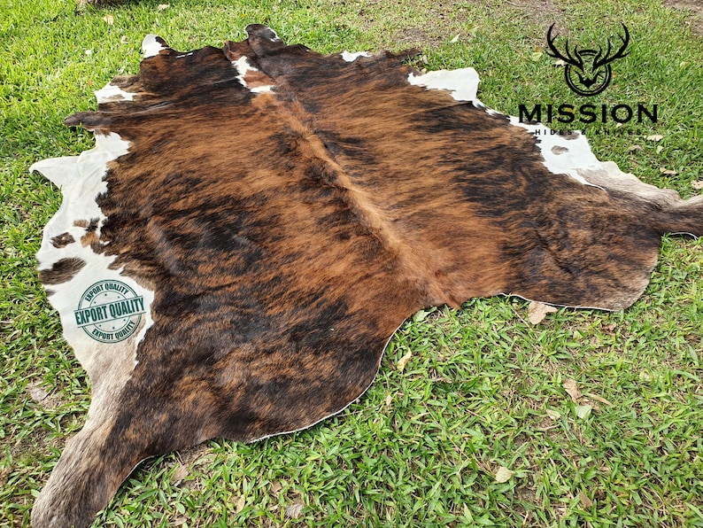 May include: A brown and white cowhide rug with a black and brown pattern. The rug is laid out on a green grassy surface. The text "EXPORT QUALITY" is printed on a white circle with a green border. The text "MISSION HIDES & LEATHERS" is printed in black on a white background.