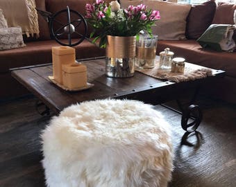 May include: A white faux fur ottoman with wooden legs sits in front of a wooden coffee table with a vase of pink flowers and candles. The coffee table is in front of a brown leather sofa with pillows.