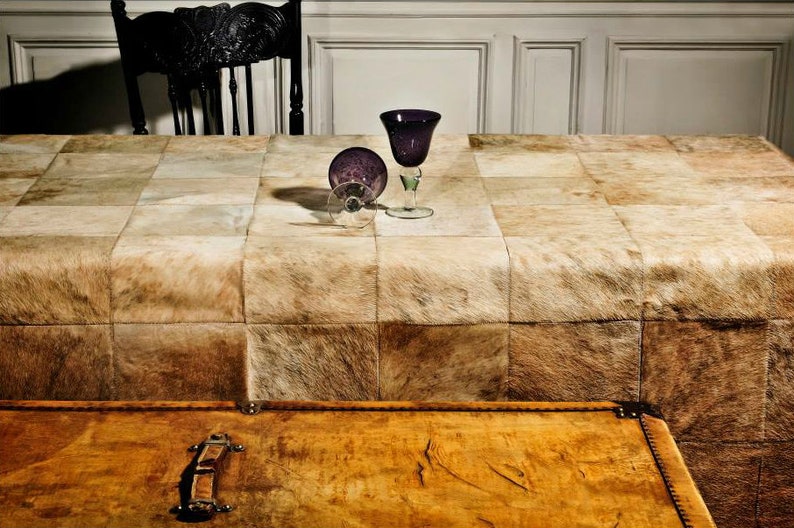 May include: A patchwork tablecloth made of light brown cowhide with two purple glass wine glasses on top.