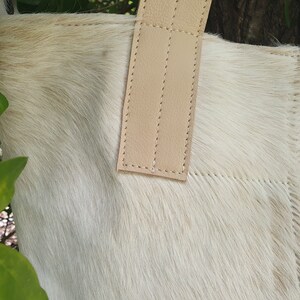 May include: A white cowhide tote bag with a light brown leather handle. The bag has a stitched design and is made of soft, natural material.