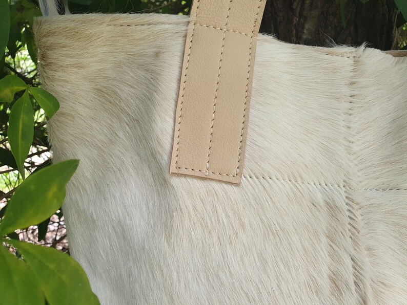 May include: A white cowhide tote bag with a light brown leather handle. The bag has a stitched design and is made of soft, natural material.