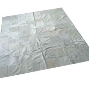 May include: A white cowhide patchwork rug with a square pattern. The rug is made of many different pieces of cowhide, each with a slightly different texture and color.