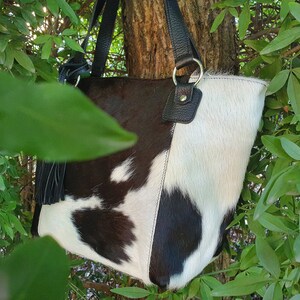 May include: A black and white cowhide tote bag with black leather handles and a black tassel. The bag is hanging from a tree branch.