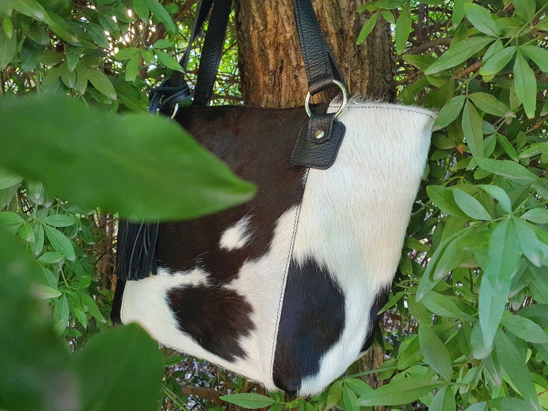 May include: A black and white cowhide tote bag with black leather handles and a black tassel. The bag is hanging from a tree branch.
