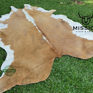 Pode incluir: Um tapete de pele de vaca marrom e branco com uma faixa branca no centro. O tapete está colocado sobre uma superfície de grama verde. O texto "MISSION HIDES & KNIT" é visível no canto superior direito da imagem. Um círculo verde com o texto "GUARANTEED 100% ORIGINAL GUARANTEED" é visível no canto inferior esquerdo da imagem.