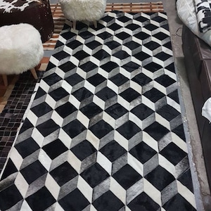 Puede incluir: Una alfombra rectangular con un diseño geométrico de cubos 3D en negro, blanco y gris. La alfombra está hecha de un material suave. Varios otomanas blancas y un otomana de piel de vaca marrón y blanca están en el fondo.