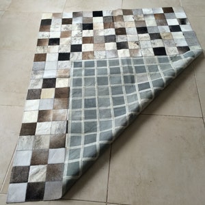 May include: A patchwork rug made of cowhide squares in various shades of brown, white, and black. The rug is folded in half, revealing a grey and white checkered pattern on the underside.