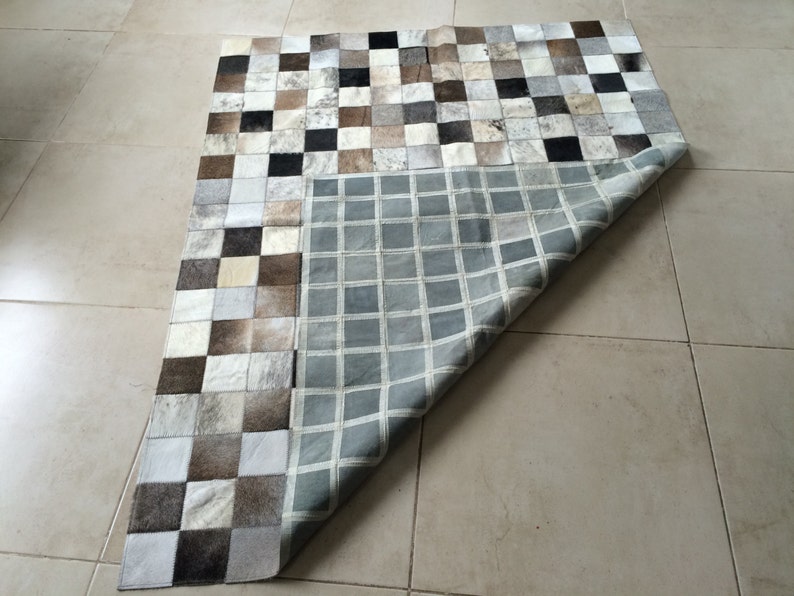 May include: A patchwork rug made of cowhide squares in various shades of brown, white, and black. The rug is folded in half, revealing a grey and white checkered pattern on the underside.