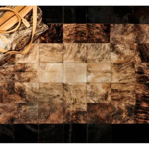 May include: A patchwork rug made of cowhide with a variety of brown and black tones. The rug is made up of squares of different sizes and shapes. A brown leather bag is lying on top of the rug.