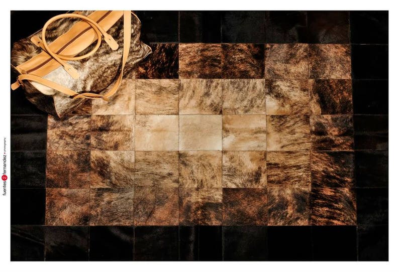 May include: A patchwork rug made of cowhide with a variety of brown and black tones. The rug is made up of squares of different sizes and shapes. A brown leather bag is lying on top of the rug.