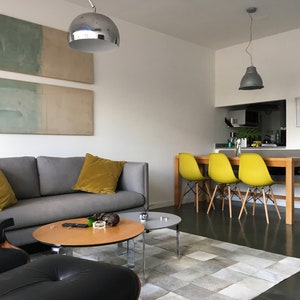 May include: A living room with a gray sofa, two yellow throw pillows, a black leather armchair, and a wooden coffee table. There is a rug on the floor and a dining area with four yellow chairs in the background.
