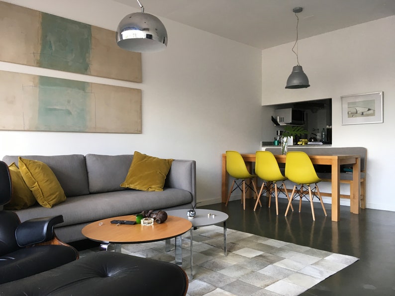 May include: A living room with a gray sofa, two yellow throw pillows, a black leather armchair, and a wooden coffee table. There is a rug on the floor and a dining area with four yellow chairs in the background.